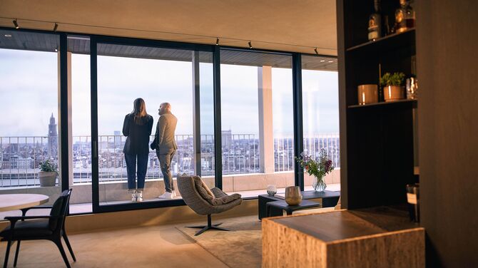 Alt: Interior view of modern apartment with wide sliding doors and two people on a balcony.