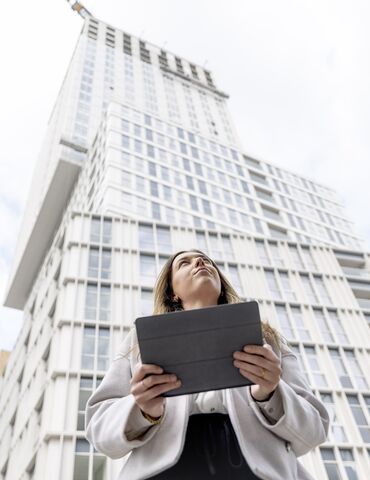 Blonde woman holding iPad looking up towards white apartment building.