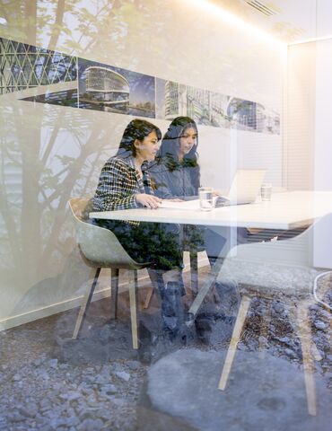 wo women sitting next to each other behind a laptop in a white meeting room.